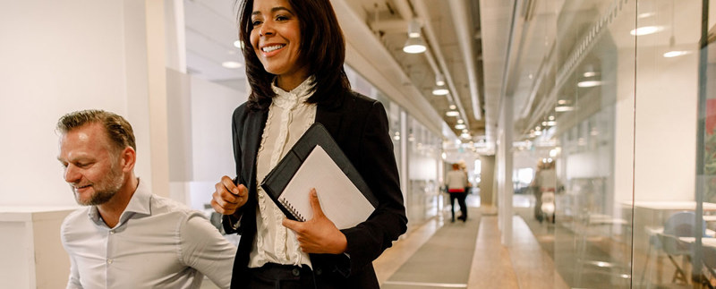 En kvinna och en man i en korridor i kontorsmiljö med anteckningsblock, pennor och laptop i handen och knät.