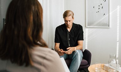 Young man in therapy  conversation with woman who can only be seen from behind. The man looks down at his hands.