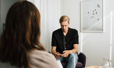 Young man in therapy  conversation with woman who can only be seen from behind. The man looks down at his hands.