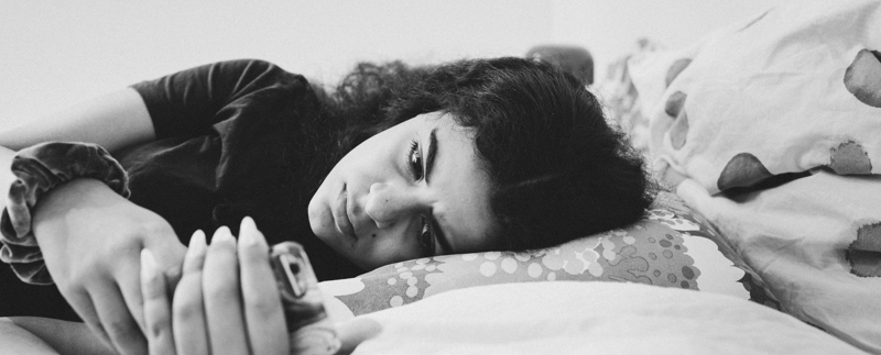 Ung kvinna ligger i en säng och läser meddelande på sin telefon med bekymrat ansiktsuttryck