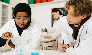 Students in white lab coats who work with liquid in test tubes.