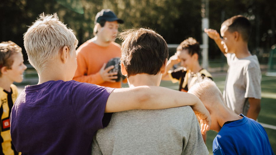 A group of boys in a soccerteam, the coach in the background. One boy has put his arm around the shoulders of another boy.